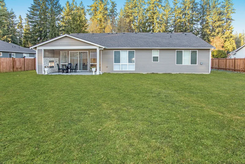 Back of the home with a large, level, fenced lawn, a covered patio and a line of evergreen trees beyond