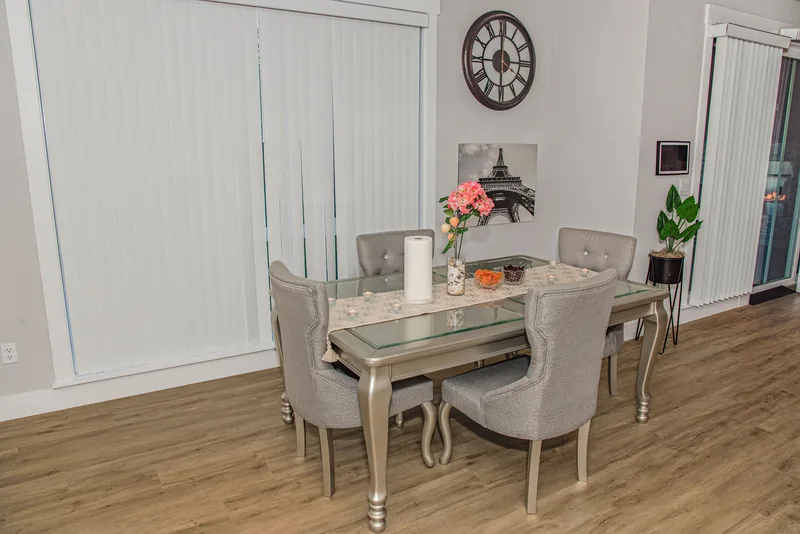 Dining area with a glass-top table, six upholstered chairs and large windows with white blinds
