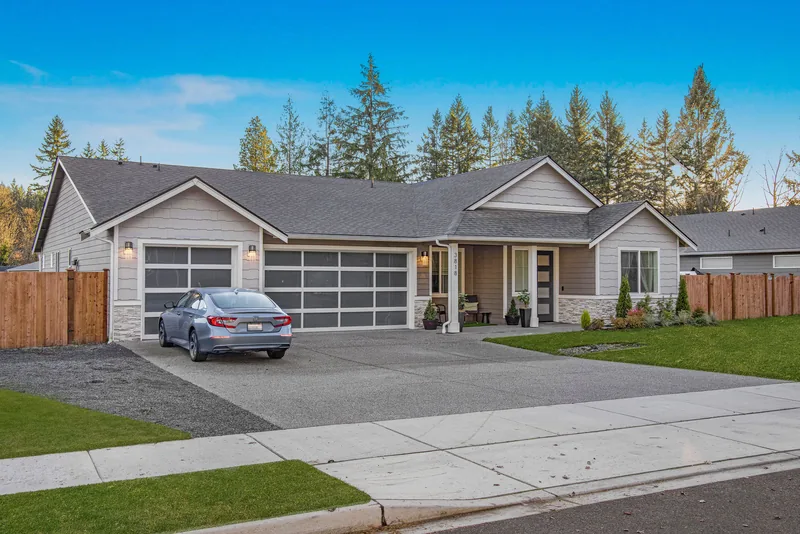 Angled daytime view of the gray single-story home with stone accents, a wide driveway, a wooden side fence and tall evergreen trees