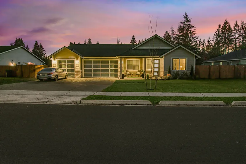 Front of the single-story gray home at dusk, with warm light glowing from the windows and frosted-glass garage doors under a pink and violet sky