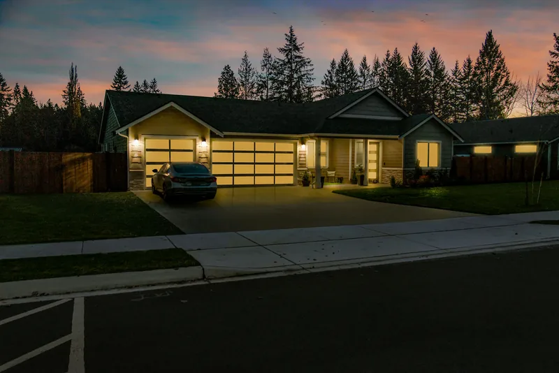 Three-quarter view of the home at twilight, with lit windows, porch lights and a glowing garage, and tall evergreens behind the roofline