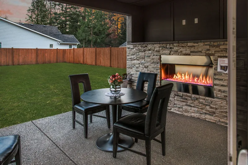 Patio at dusk with the outdoor fireplace lit, a round table with a small vase of flowers, and the fenced lawn beyond